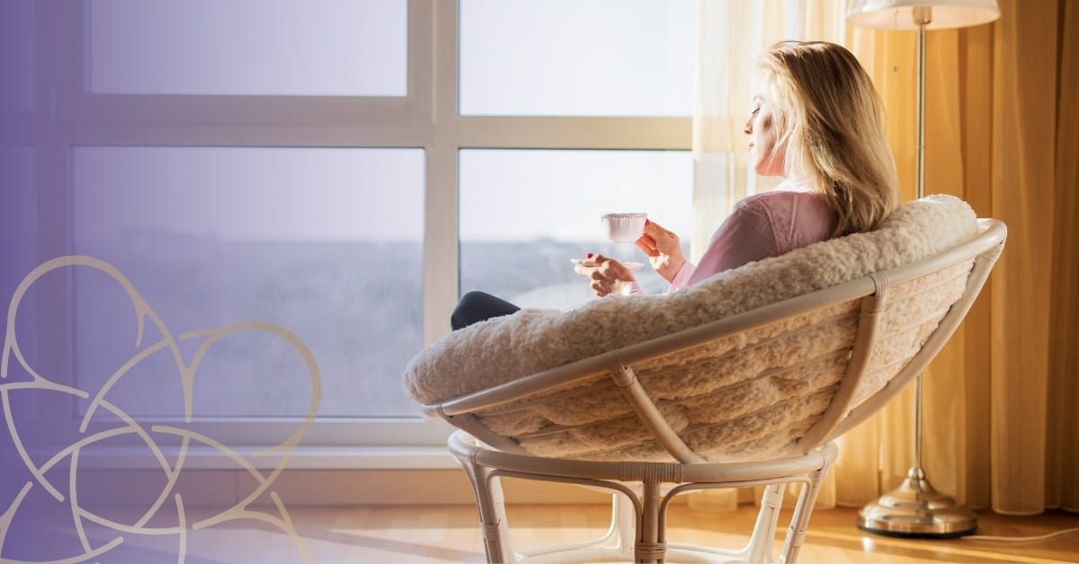 A woman sits in a cosy round chair by a large bright window, holding a cup and saucer, bathed in warm sunlight. Curtains are drawn back, a lamp stands beside her—she reflects on personal growth clarity as a decorative pattern overlays the left side.