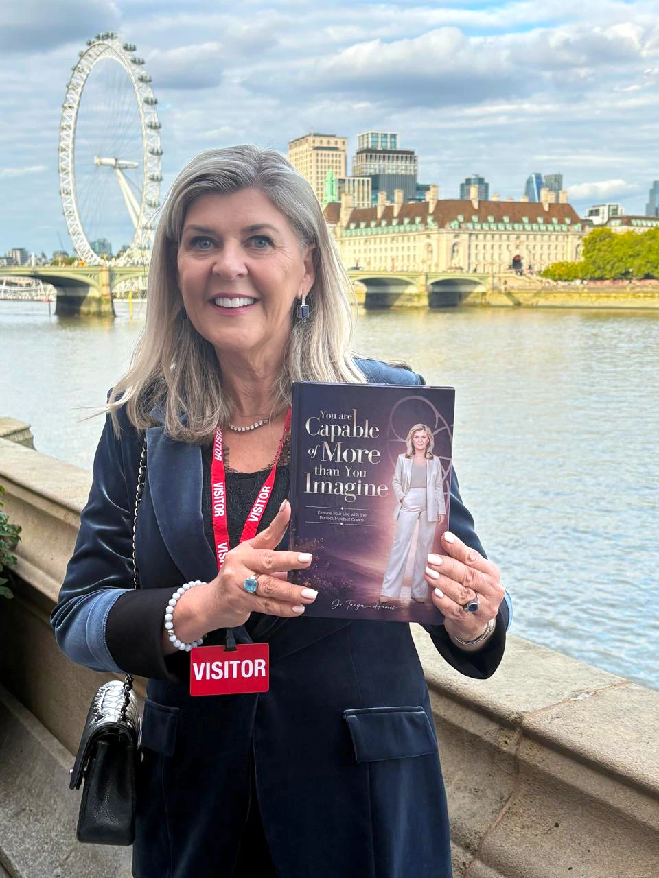 Dr Tanya Hames holding her book “You Are Capable of More Than You Imagine” beside the River Thames in London, with the London Eye in the background.